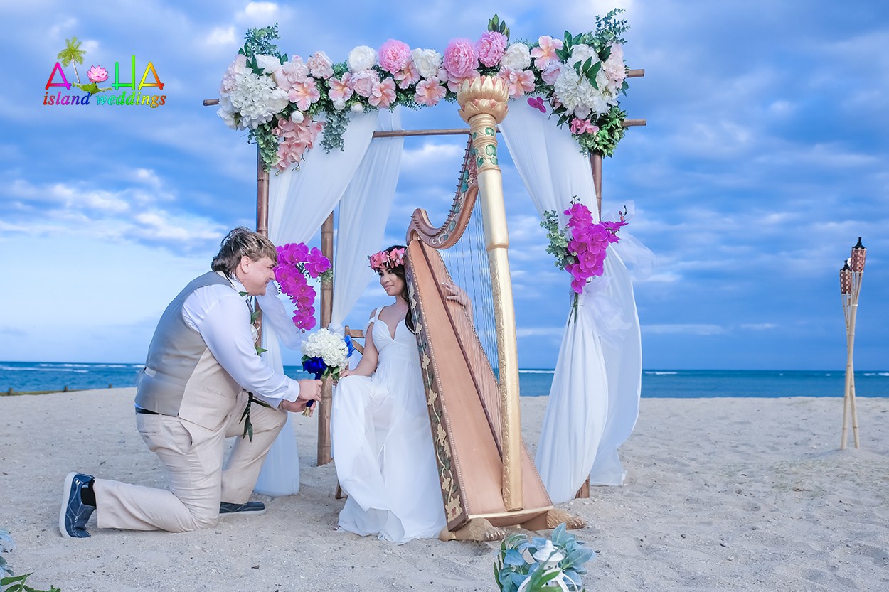 Groom kneeling on the sand to hand a white and blue bouquet to his bride seated at a golden harp under a pink flower arch by the ocean